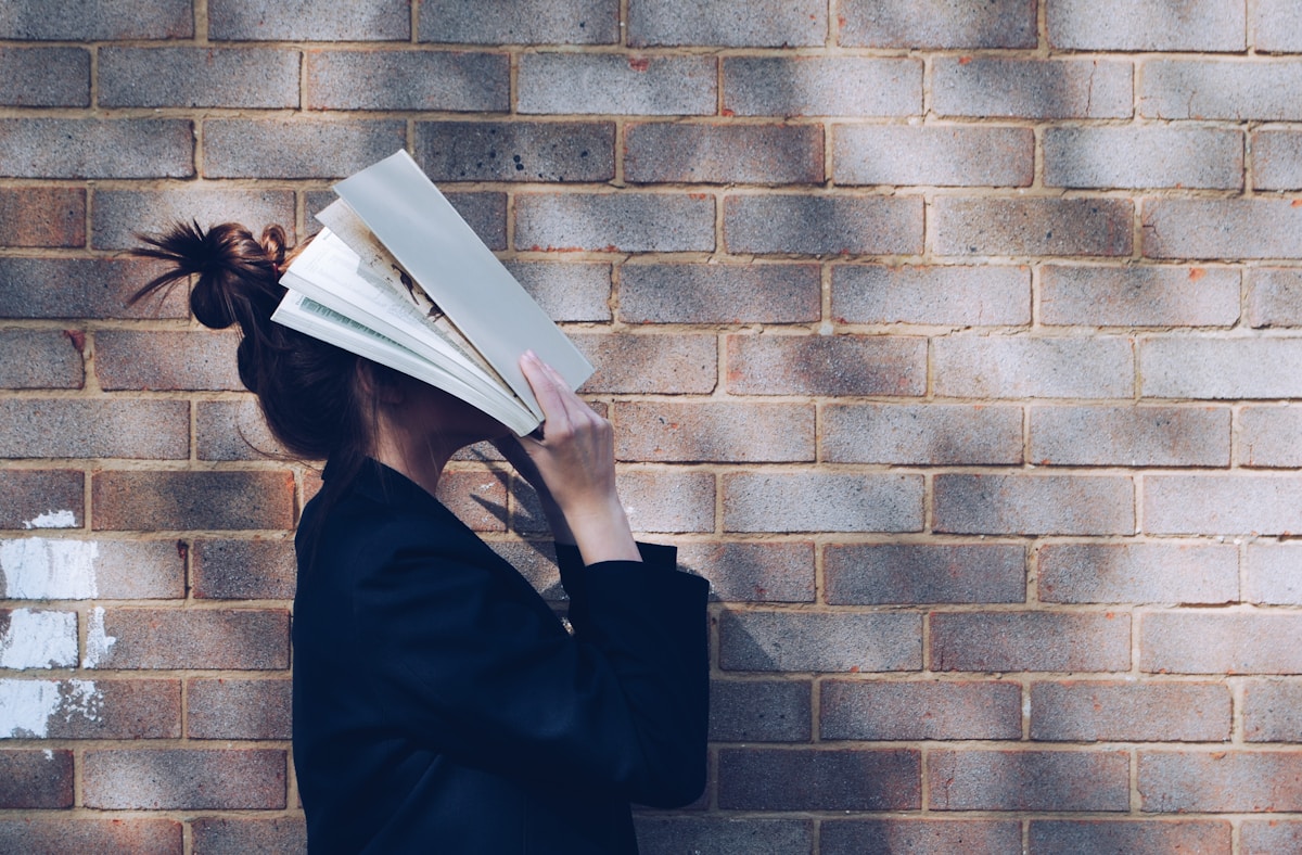 Student hiding their face behind an open book against a brick wall